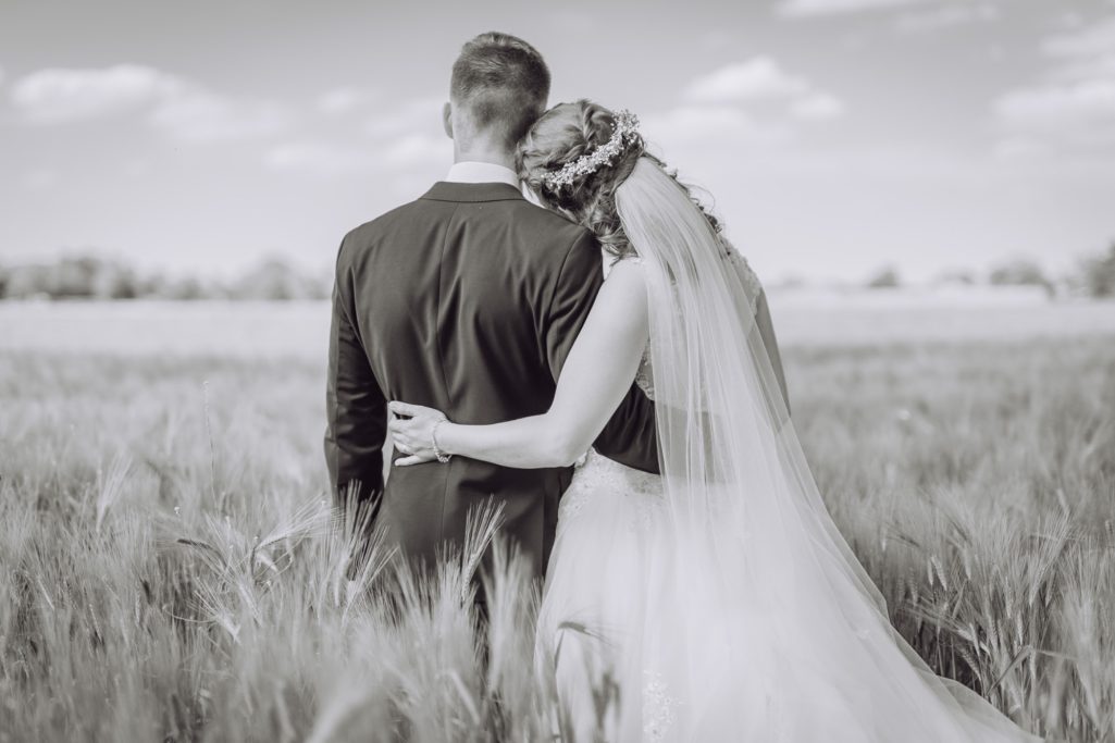 Ein Brautpaar steht in einem Feld, umarmt sich romantisch, im Hintergrund Himmel und Wolken.