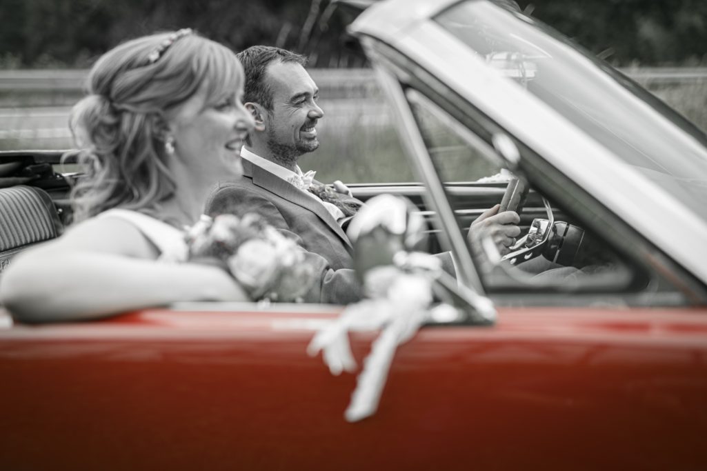 Braut und Bräutigam sitzen lächelnd in einem roten Cabrio mit Blumen.
