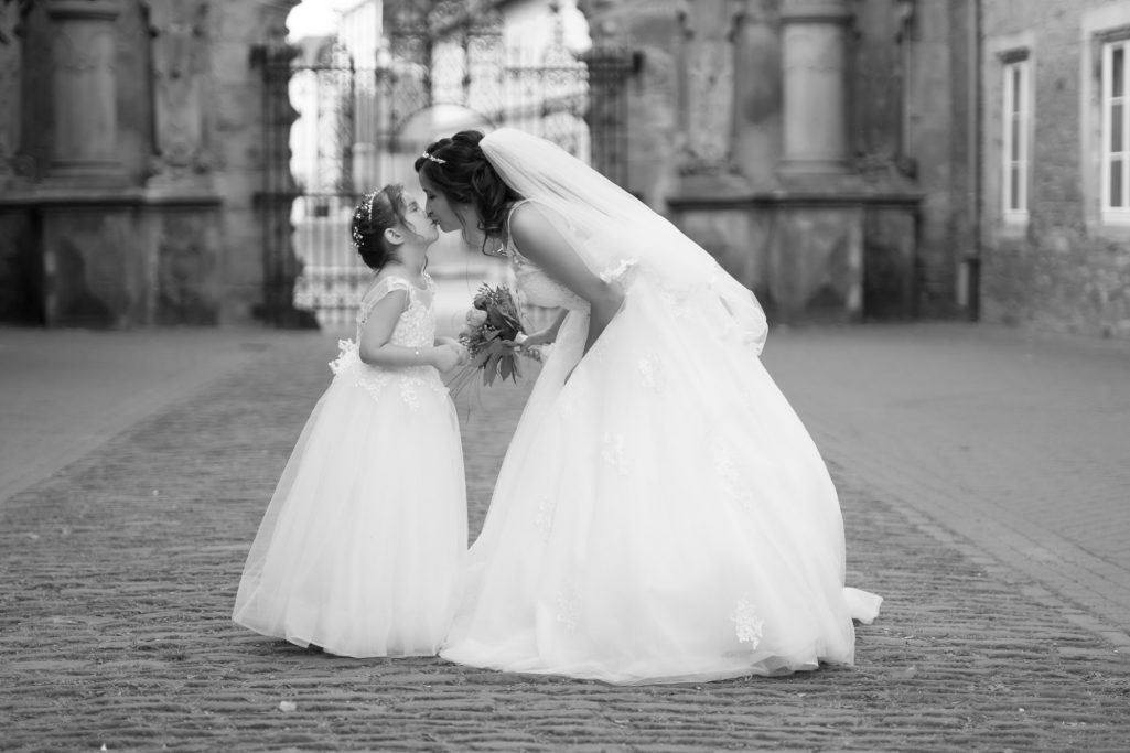 Braut in weißem Kleid beugt sich zu kleinem Mädchen in ähnlichem Kleid, beide lächeln.