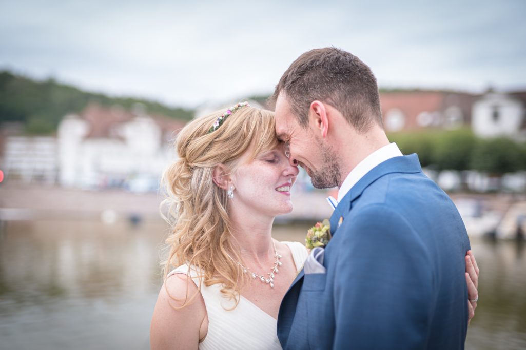 Hochzeitspaar in Zärtlichkeit umarmt, mit Blick auf Wasser und Stadt im Hintergrund.