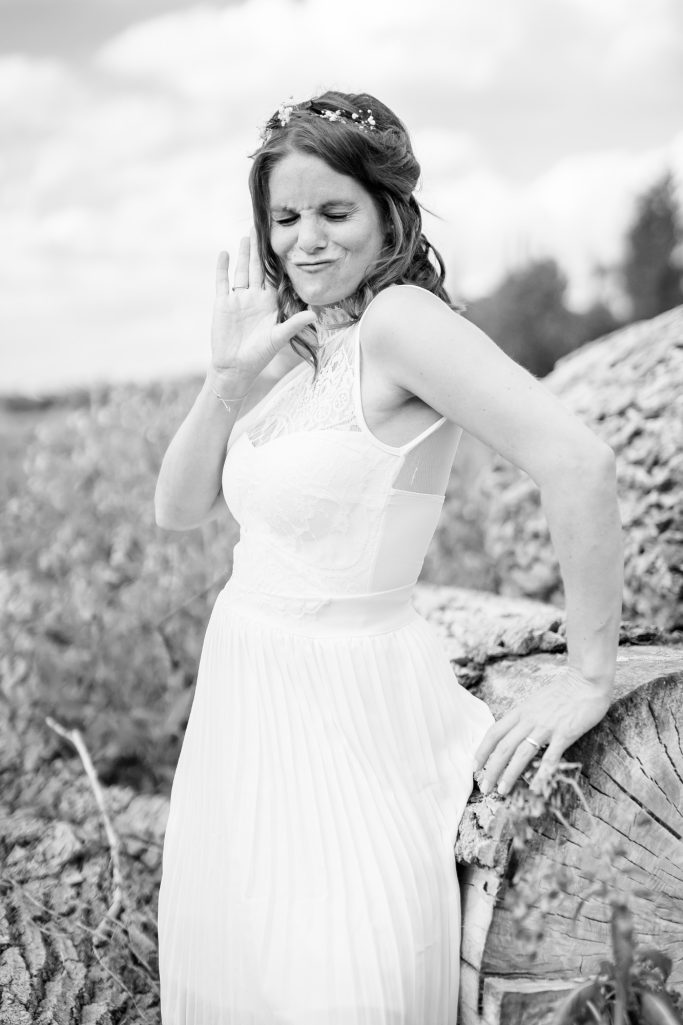 Frau in einem weißen Kleid steht lächelnd neben einem Baumstamm im Freien.