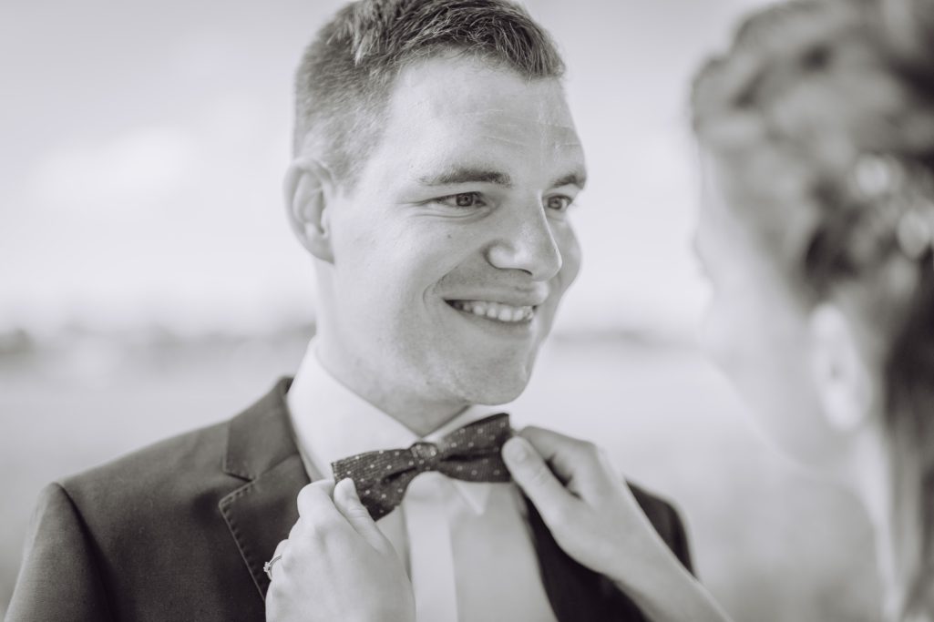 Brautpaar, Bräutigam trägt einen Anzug und lächelt, während die Braut seine Fliege richtet.