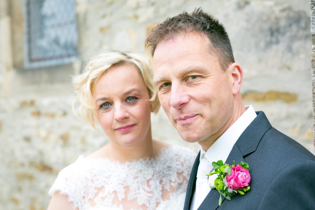 Hochzeitsfoto eines Paares vor einer steinernen Wand, Braut mit weißem Kleid und Rosenstrauß.