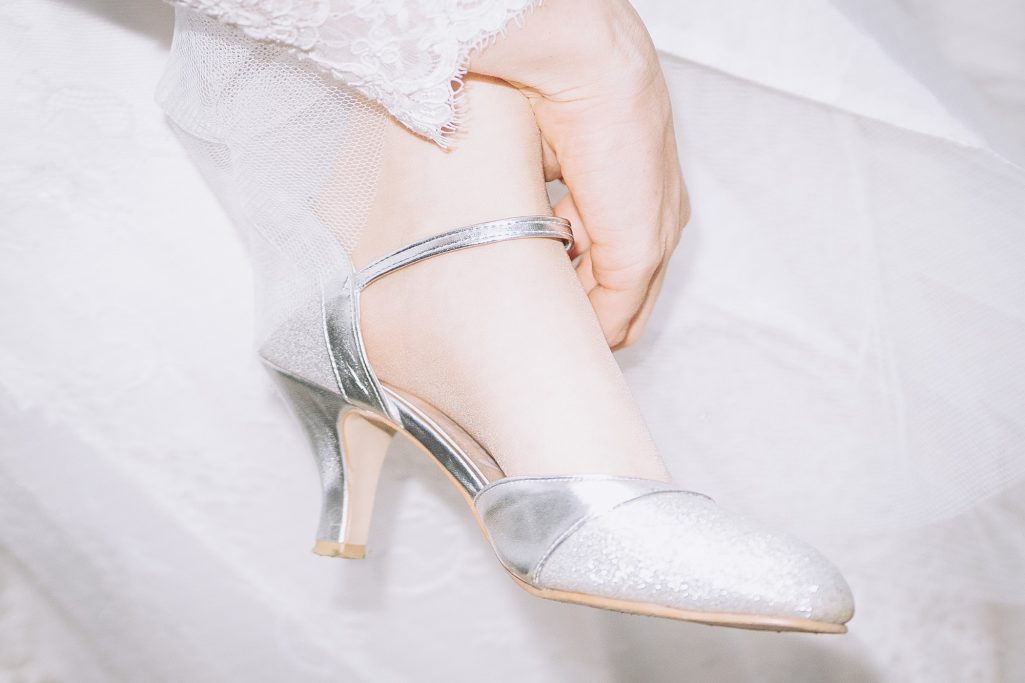 Eleganter silberner High Heel mit dünnem Absatz, an einem Fuß in Hochzeitskleid.