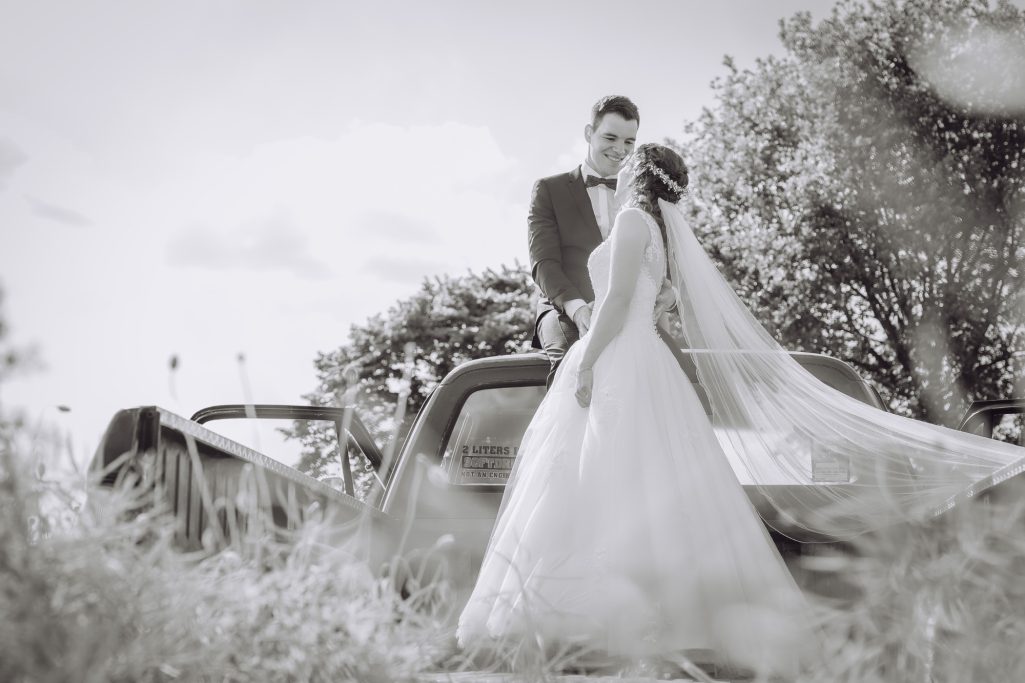 Braut und Bräutigam stehen romantisch vor einem Oldtimer in der Natur.