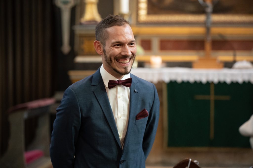 Lächelnder Mann in Anzug mit Fliege vor einer kirchlichen Kulisse.