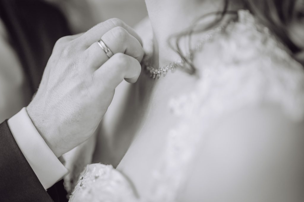 Eine Hand befestigt eine Kette am Hals einer Braut in einem eleganten Kleid.