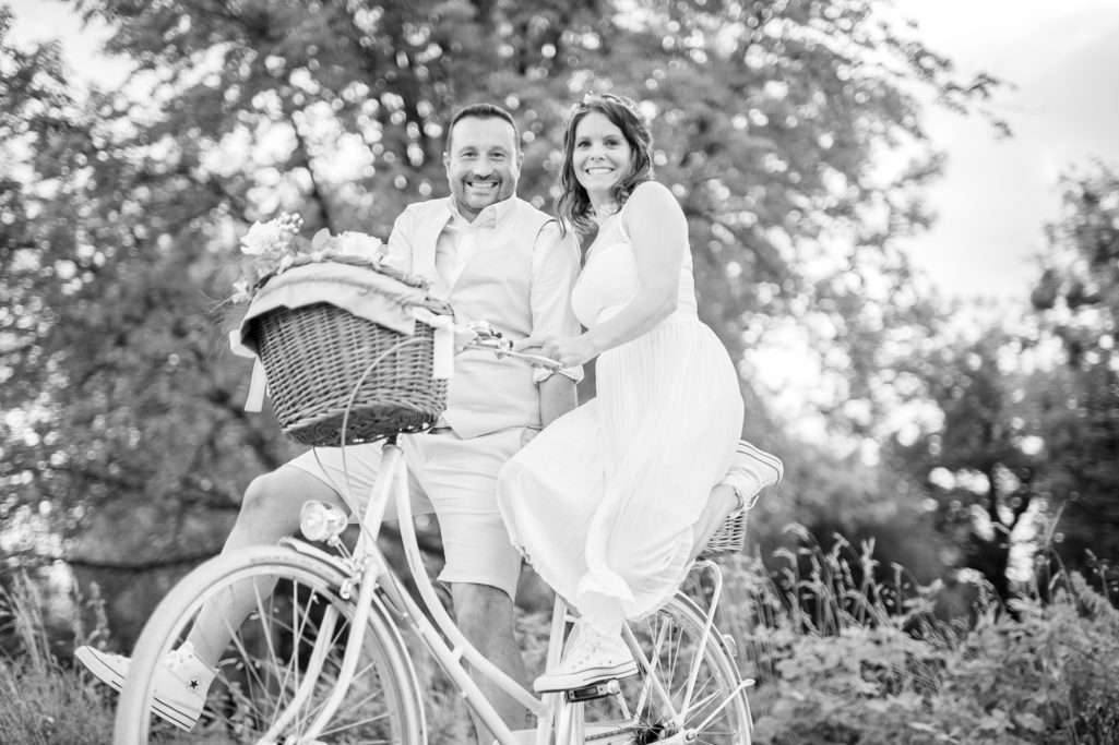 Ein Paar sitzt auf einem Fahrrad mit einem Korb, umgeben von grünen Bäumen.