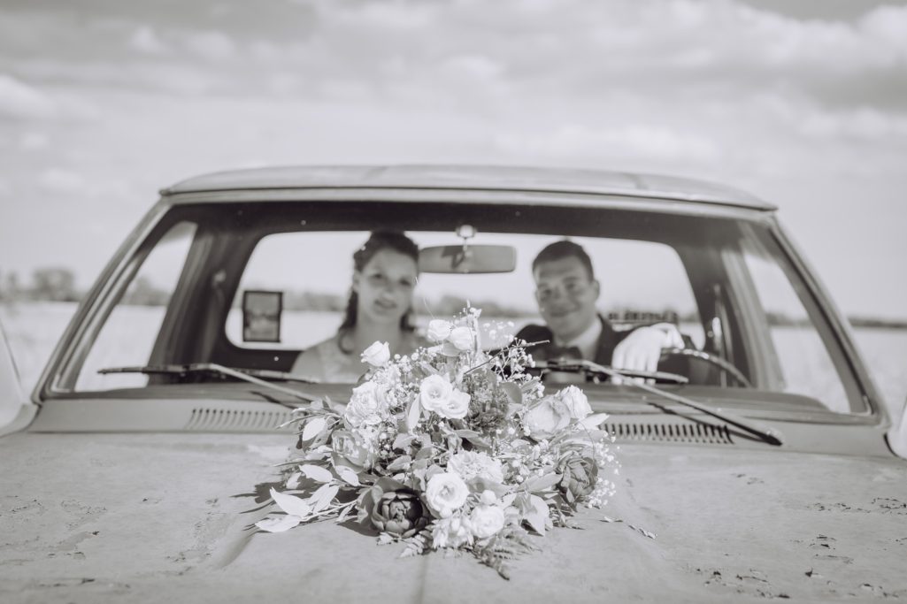 Ein Paar sitzt im Auto, lächelnd, mit einem Blumenstrauß auf dem Armaturenbrett.