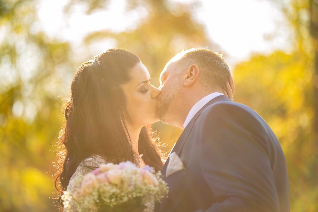 Braut und Bräutigam küssen sich romantisch im Sonnenlicht mit Blumenstrauß.