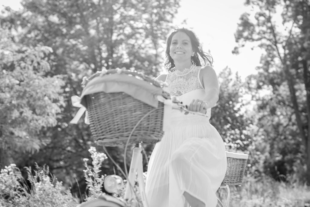 Frau in weißem Kleid fährt fröhlich auf einem Fahrrad mit einem Korb durch die Natur.