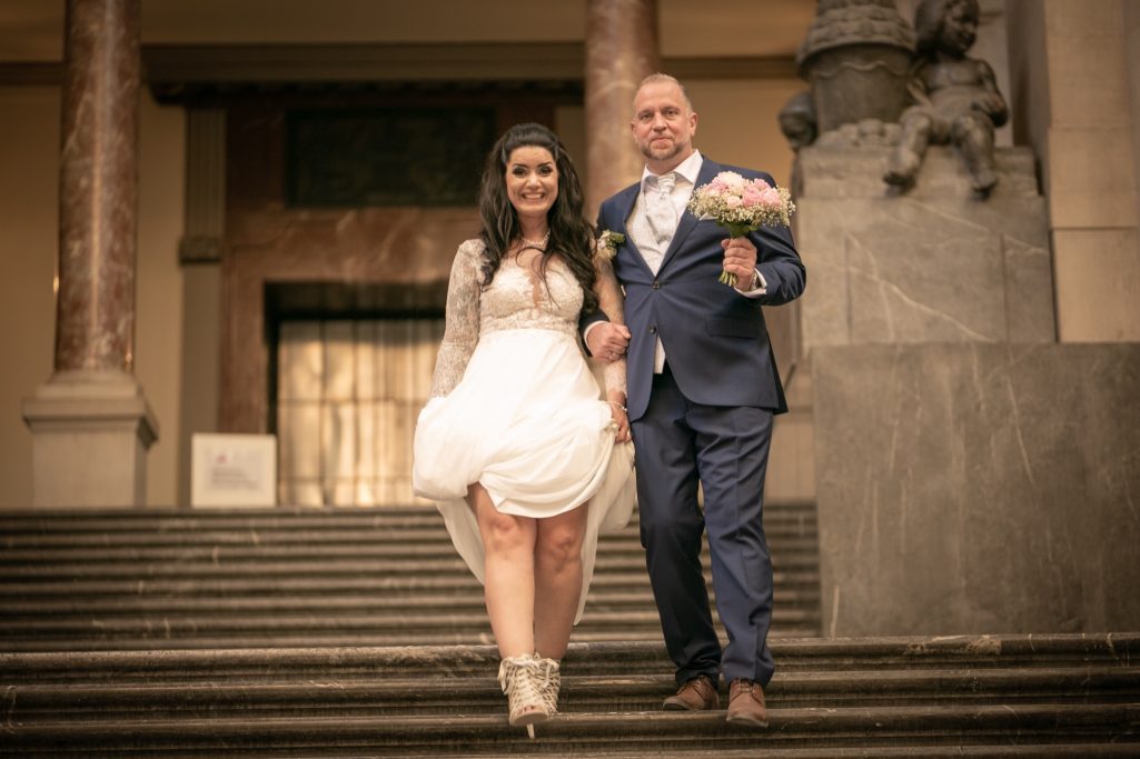 Braut und Bräutigam gehen eine Treppe hinunter, beide elegant gekleidet.