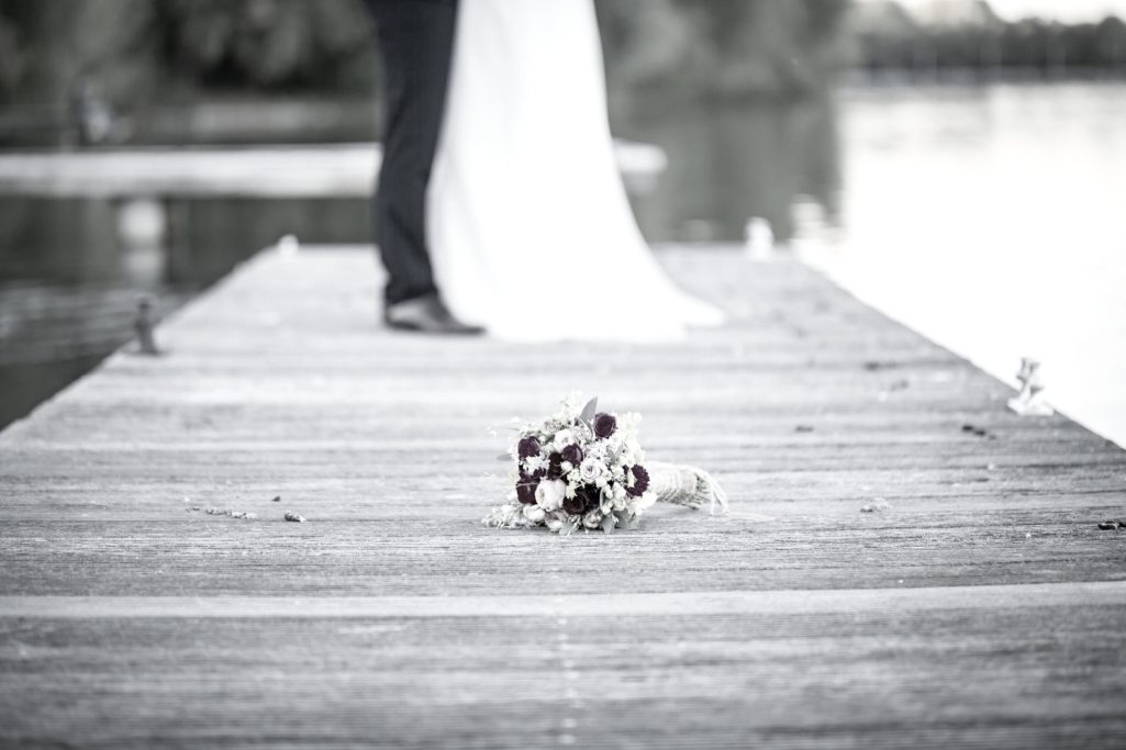 Brautstrauß auf einem Steg, im Hintergrund ein Paar in Hochzeitskleidung.